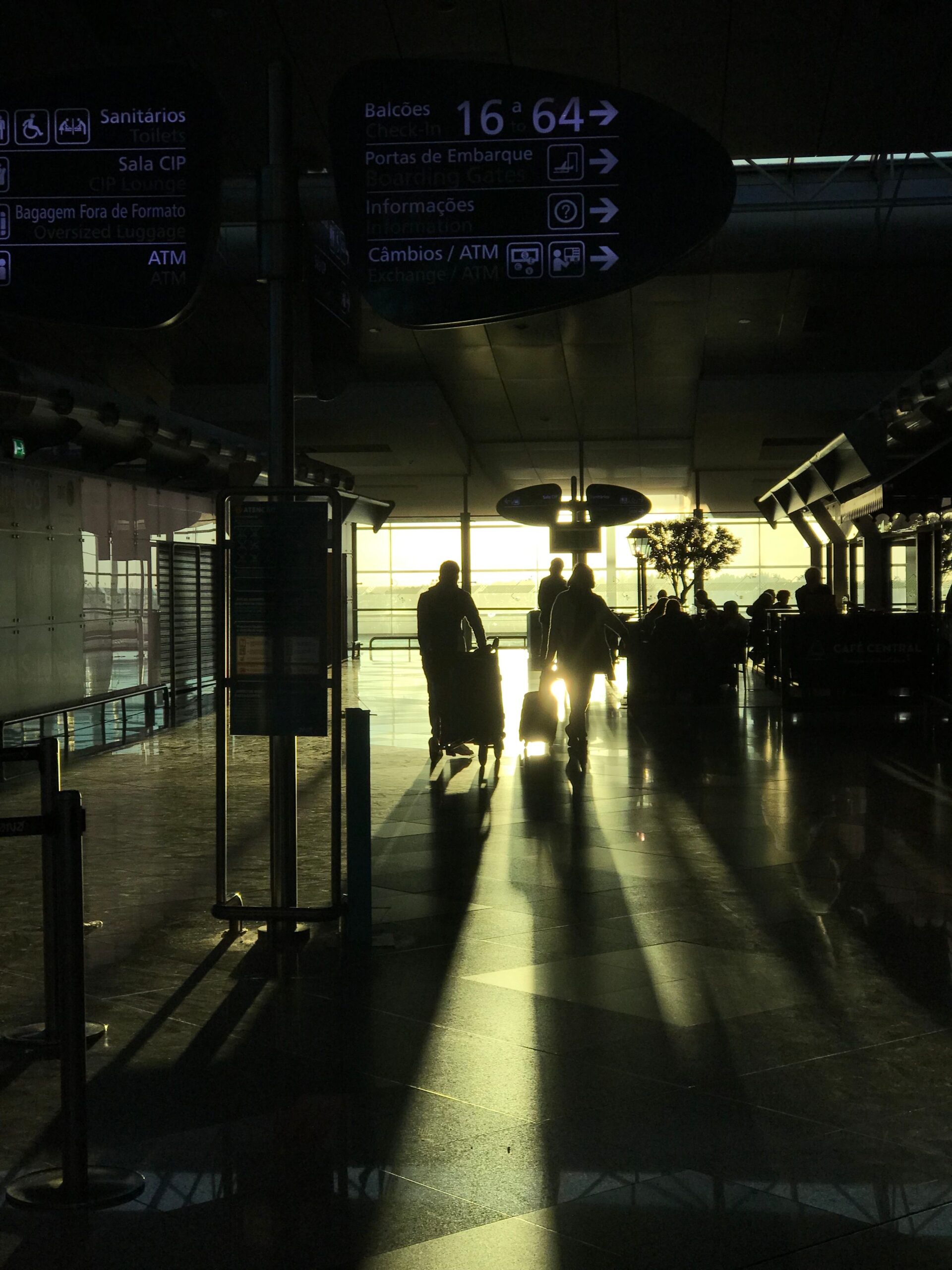 Terminal de aeroporto europeu ao entardecer: viajantes em silhueta caminham com malas em direção à luz dourada que entra pelas grandes janelas envidraçadas; à direita, uma área de café e uma árvore.