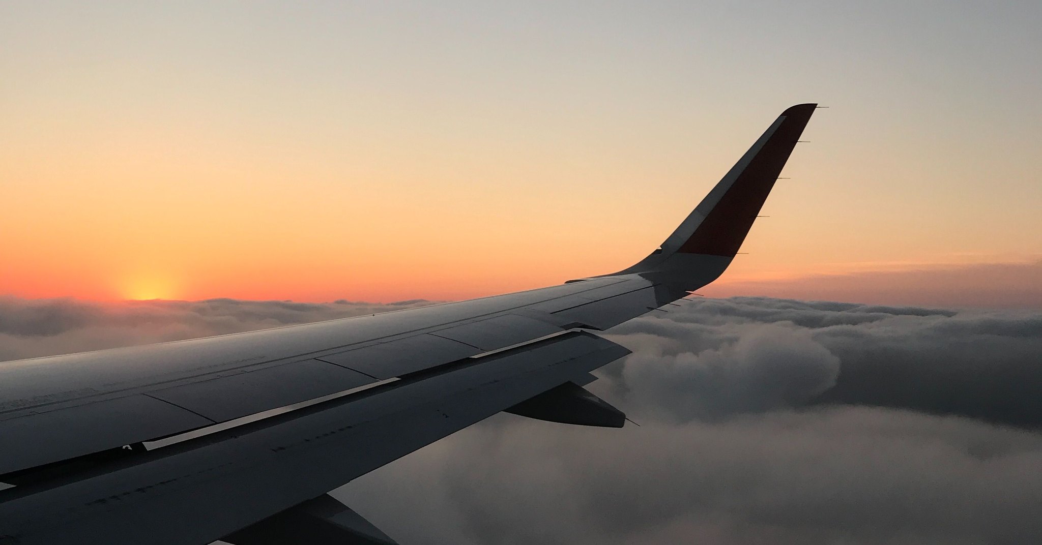 Asa de um avião vista da janela durante o pôr do sol, acima de uma camada de nuvens.