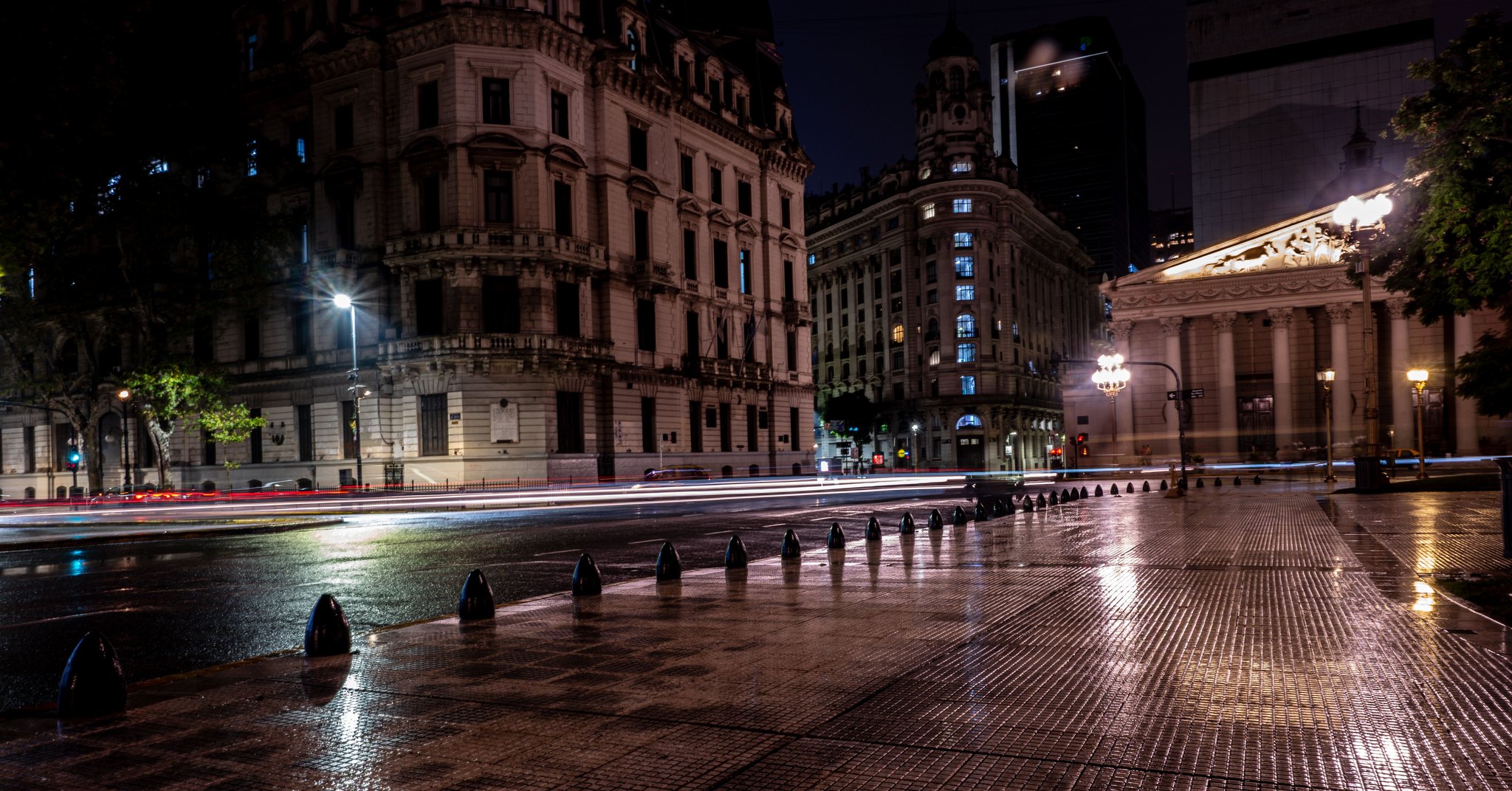 Rua de Buenos Aires à noite, com prédios históricos iluminados, calçada molhada refletindo as luzes e rastros de faróis atravessando a avenida.
