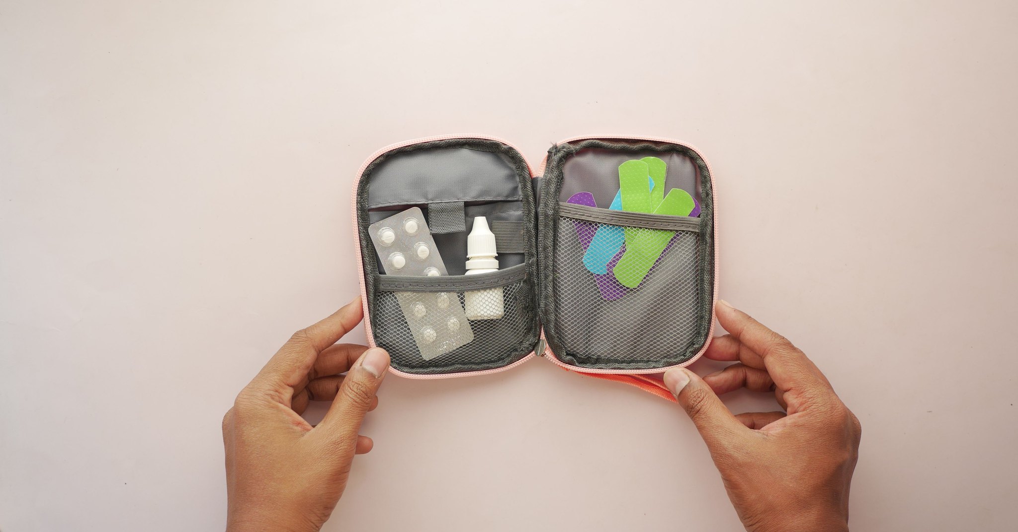 Duas mãos segurando um estojo de primeiros socorros aberto, com cartelas de comprimidos, um frasco branco sem rótulo e curativos coloridos nos compartimentos de tela.