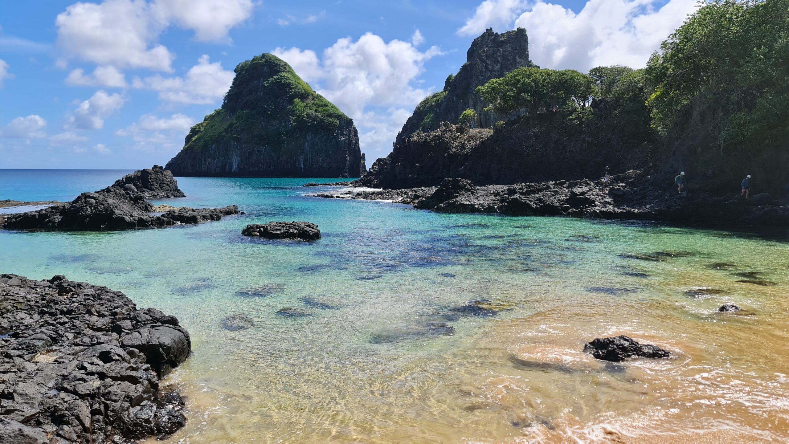 Vista da Baía dos Porcos, em Fernando de Noronha, com o Morro Dois Irmãos ao fundo e água transparente sobre as pedras.