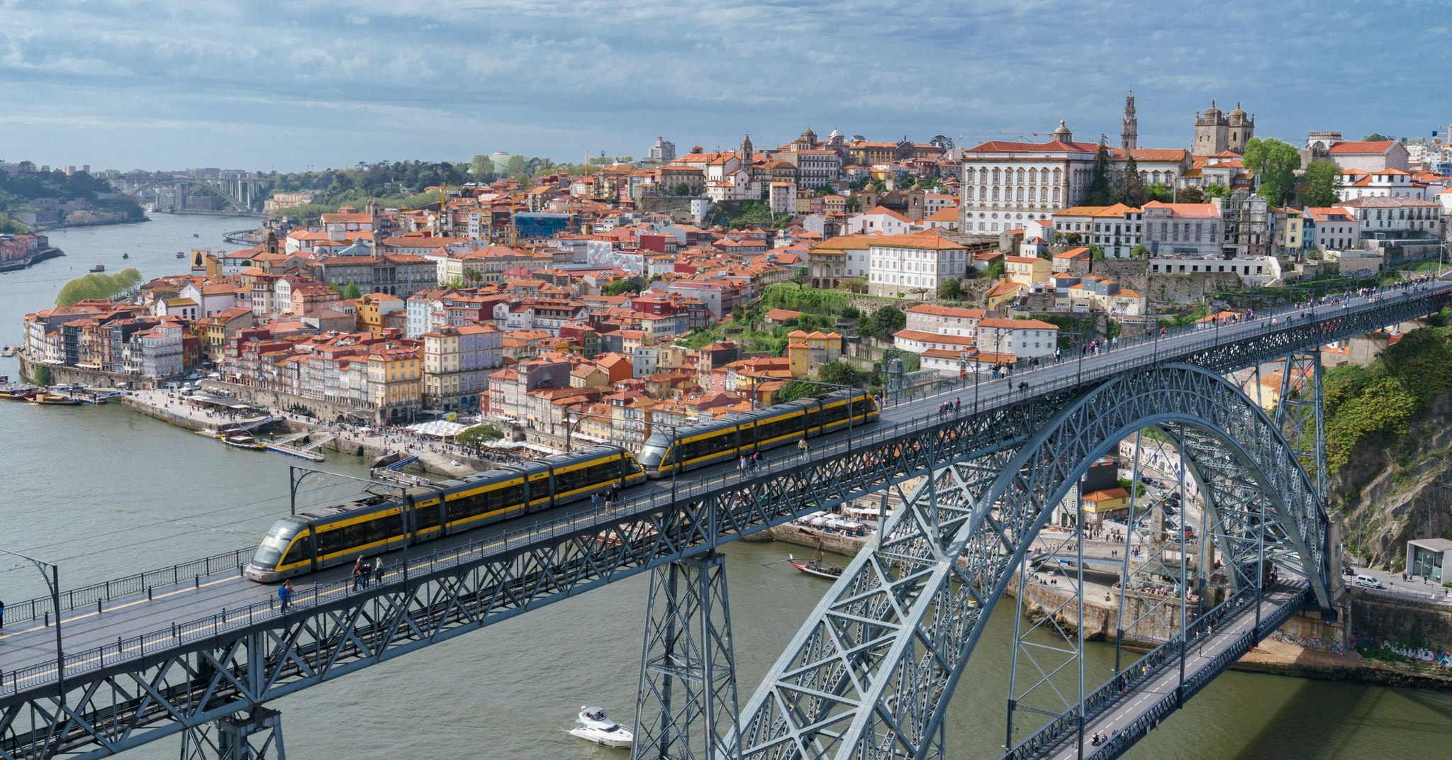Vista aérea da Ponte Luís I, no Porto, com o metro atravessando o tabuleiro superior, o rio Douro embaixo e as casas de telhado laranja da Ribeira ao fundo.