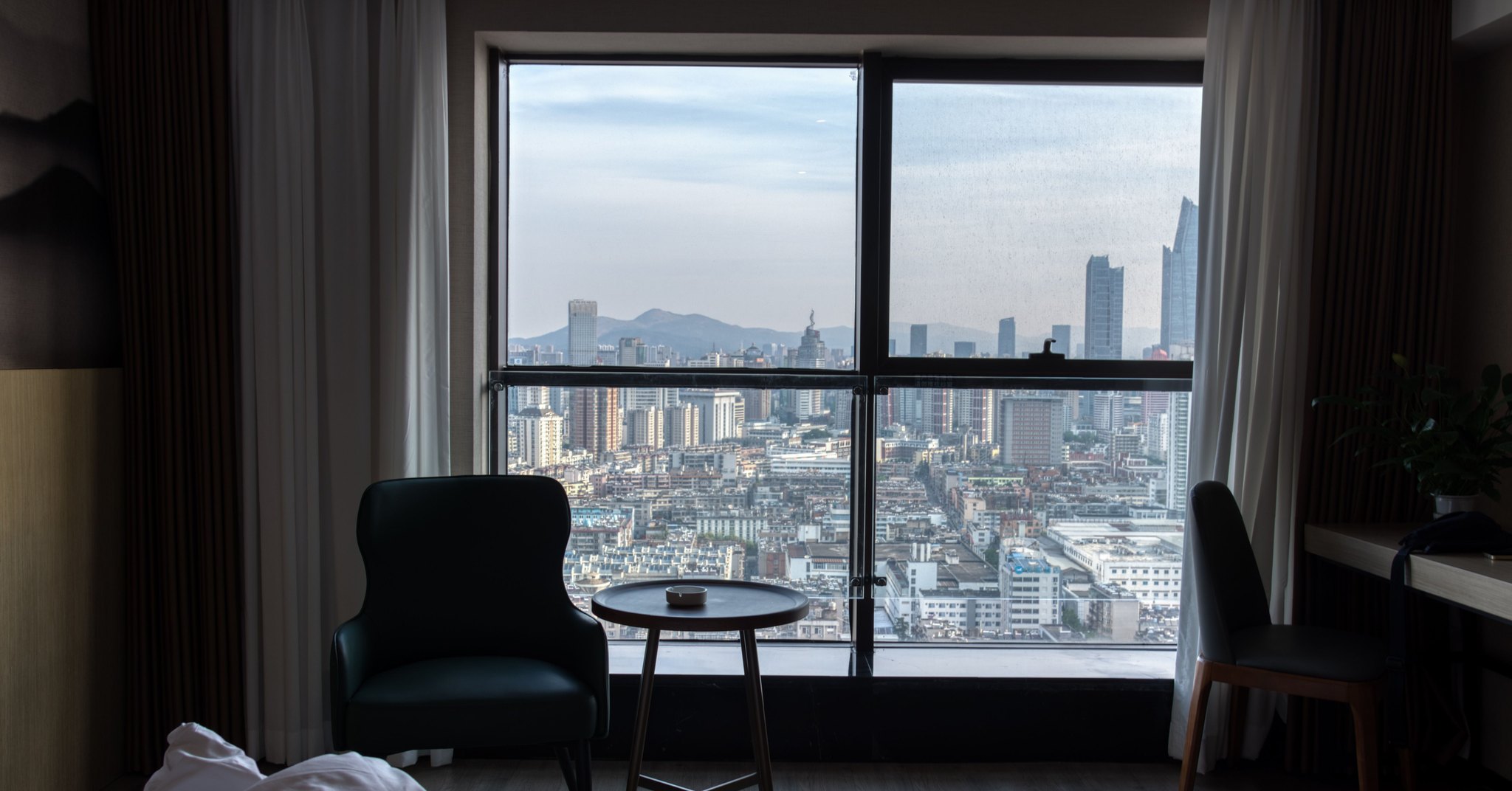 Interior de quarto de hotel com poltrona, mesa de apoio e janela ampla que ocupa a parede, com vista para prédios de uma cidade ao amanhecer.