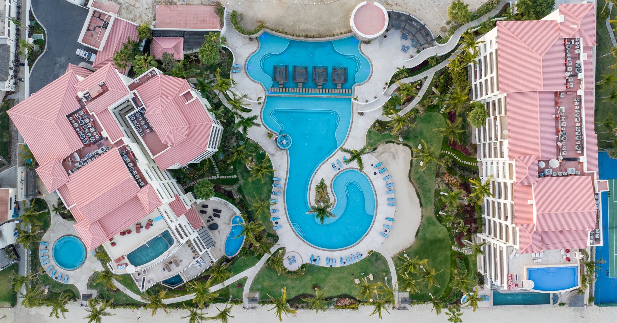 Vista aérea a prumo de um complexo de resort na beira do mar, com piscinas de formato irregular, coqueiros, prédios de telhado vermelho e a faixa de areia na parte de baixo.