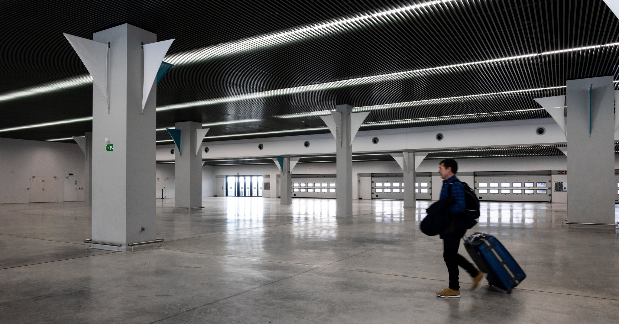 Saguão amplo e quase vazio de aeroporto, com colunas claras e forro escuro iluminado por faixas de luz; à direita, um viajante caminha puxando uma mala de rodinhas.